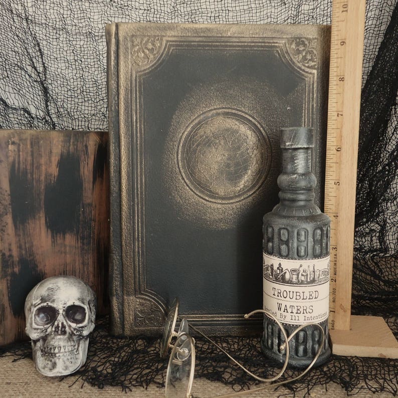 May include: A still life arrangement featuring a skull, antique book, and a bottle labeled "Troubled Waters." The book is black with gold accents. The bottle is gray with a label. A pair of round glasses rests in front of the bottle. A wooden ruler is in the background.