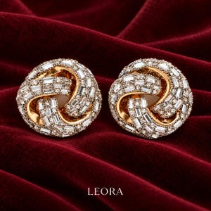 May include: A pair of gold-toned stud earrings featuring a circular, knotted design encrusted with numerous clear baguette and round-cut stones. The word LEORA is printed in white capital letters at the bottom of the image.