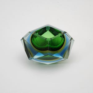 Puede incluir: Un cuenco de cristal facetado con un núcleo interior de color verde esmeralda intenso encerrado en capas de cristal transparente y azul. El objeto tiene una forma exterior geométrica de múltiples lados con bordes afilados y un acabado pulido.