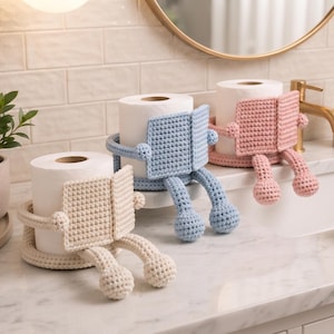 May include: Three crochet toilet paper holders shaped like figures reading books. Each holder features a circular base, two dangling legs with rounded feet, and a rectangular book-shaped front panel. The holders are in cream, light blue, and pink.
