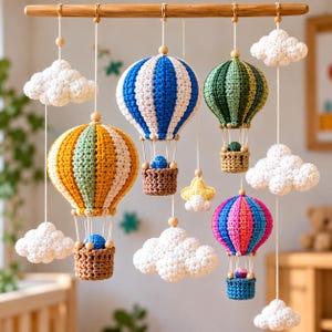 Puede incluir: Un móvil colgante con cuatro globos aerostáticos de ganchillo a rayas en azul y blanco, amarillo y verde, verde y verde oscuro, y rosa y azul. El móvil incluye nubes blancas de ganchillo y una pequeña estrella amarilla de una barra de madera.