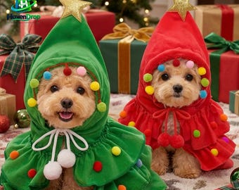 Puede incluir: Dos perros pequeños con capas festivas con capucha. Una capa es verde y la otra roja, ambas con una estrella dorada en la parte superior, volantes escalonados y pompones de colores. Las capas incluyen cordones blancos con pompones.