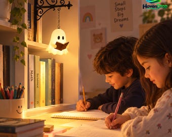 Puede incluir: Una luz nocturna blanca con forma de fantasma que cuelga de un soporte de pared de metal negro. La figura del fantasma está iluminada, emite un brillo cálido y sostiene un pequeño libro marrón. La luz cuelga de una cadena corta del soporte.