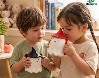 Puede incluir: Dos tazas de cerámica con forma de fantasma y tapa. Una taza tiene una tapa negra y la otra una tapa roja. Ambas tazas son blancas y tienen un rostro sencillo con dos ojos pequeños y una boca. Las tazas tienen un asa en el lateral.