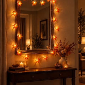 May include: A string of LED lights featuring orange and red maple leaf covers draped around a rectangular mirror with a dark wood frame. The lights emit a warm glow, illuminating the mirror and the surrounding wall with autumn-inspired colors.