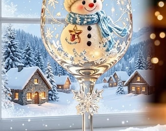 Puede incluir: Una copa de vino transparente con una figura decorativa de muñeco de nieve en el interior, que lleva un gorro y una bufanda de punto azul. El tallo tiene un adorno de copo de nieve plateado y la base incluye el texto Winter Joy.