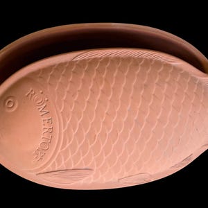May include: A terracotta fish-shaped baking dish with a lid featuring embossed scale patterns and fins. The word ROMERTOPF is clearly molded into the side of the lid. The clay material has a matte, natural reddish-brown finish.