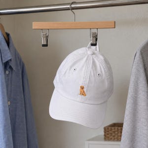 May include: A white baseball cap featuring a small, embroidered golden retriever dog sitting in the centre of the front panel. The cap has a curved brim, a button on the top, and stitched eyelets for ventilation, suspended from a wooden hanger.