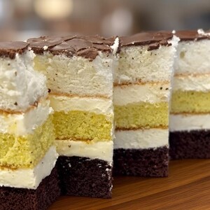 Puede incluir: Cuatro rebanadas rectangulares de pastel en capas dispuestas en fila. Cada rebanada presenta una base de chocolate oscuro, una capa de crema blanca, una capa de bizcocho amarillo, otra capa de crema blanca, una cobertura blanca y un glaseado.
