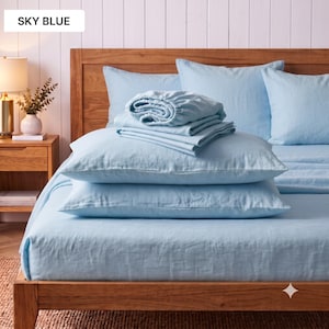 May include: A set of sky blue bed linen including a fitted sheet, a flat sheet, and two pillowcases. The bedding is arranged on a wooden bed frame, featuring a smooth, solid light blue colour throughout the entire textile collection.
