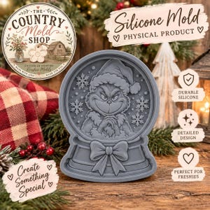 May include: A gray silicone mold shaped like a snow globe featuring a central character with a furrowed brow and a Santa hat, surrounded by snowflakes and a bow at the base. The mold has a smooth, matte finish and a raised, detailed design.