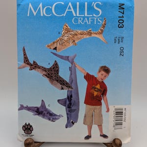 May include: A McCall's Crafts sewing pattern envelope M7103 for shark plush toys. The light blue package features illustrations of three different shark designs labeled A, B, and C, along with a photo of a child holding a large gray shark plush.