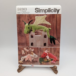 May include: A Simplicity S9363 sewing pattern envelope featuring illustrations of three plush dragon toys. The top image shows a green dragon on a castle, while the bottom images display a white dragon with pink wings and a red dragon with gold coins.
