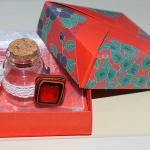 Op de afbeelding: Een klein doorzichtig glazen flesje met een kurken stop en een wit gehaakt bandje, vergezeld van een rechthoekige ring met een rood midden en gelaagde zwarte en oranje randen, gepresenteerd in een rode doos met een bloemrijk deksel.