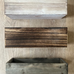 May include: Three rectangular wooden boxes in varying shades of brown and gray. The top box is light gray, the middle box is dark brown, and the bottom box is a muted gray. The boxes are empty and have a rustic, weathered appearance.