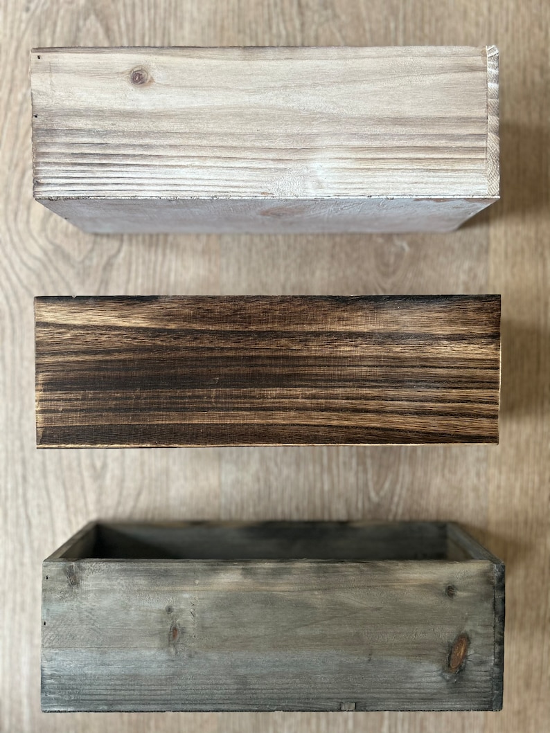 May include: Three rectangular wooden boxes in varying shades of brown and gray. The top box is light gray, the middle box is dark brown, and the bottom box is a muted gray. The boxes are empty and have a rustic, weathered appearance.