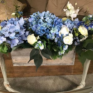 May include: A floral arrangement featuring blue hydrangeas, white and yellow flowers, and green foliage in a rustic wooden box. The arrangement sits on a glass-topped table with light-colored legs, creating a fresh and inviting centerpiece.