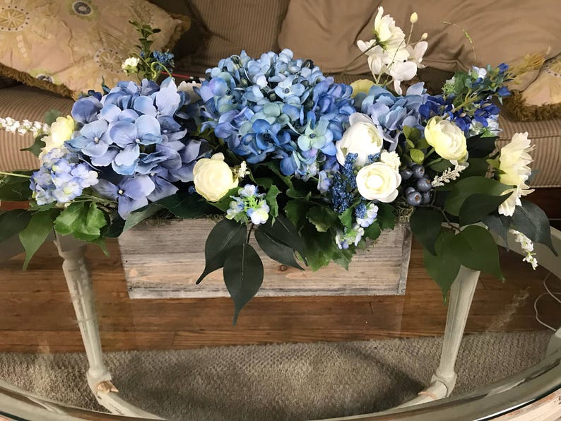 May include: A floral arrangement featuring blue hydrangeas, white and yellow flowers, and green foliage in a rustic wooden box. The arrangement sits on a glass-topped table with light-colored legs, creating a fresh and inviting centerpiece.