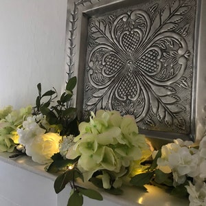 May include: A white fireplace mantel decorated with a garland of white and green flowers and lights.
