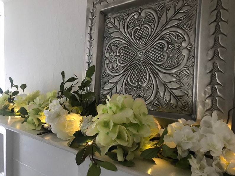 May include: A white fireplace mantel decorated with a garland of white and green flowers and lights.