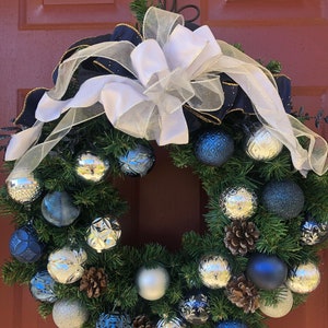 May include: A Christmas wreath with a white and navy blue ribbon bow. The wreath is decorated with silver and blue ornaments, pine cones, and greenery.