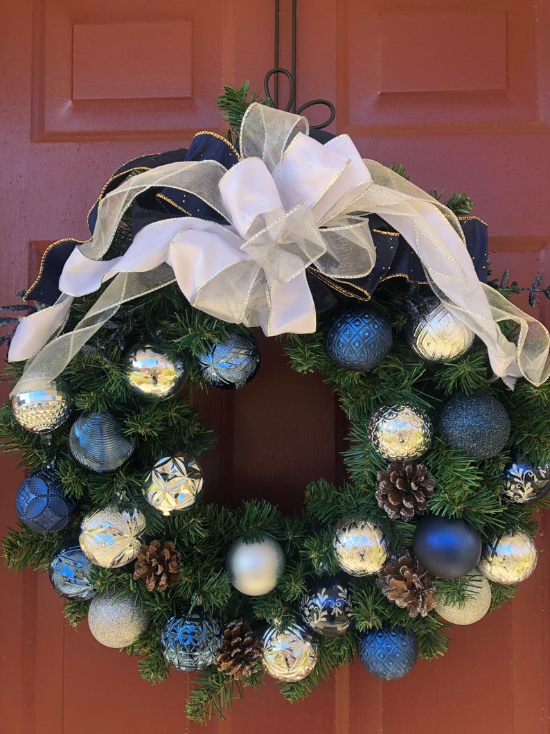 May include: A Christmas wreath with a white and navy blue ribbon bow. The wreath is decorated with silver and blue ornaments, pine cones, and greenery.