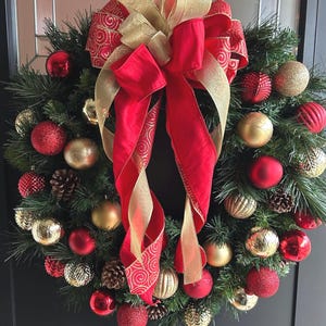 May include: A Christmas wreath with a red and gold bow, decorated with red and gold ornaments. The wreath is made of evergreen branches and has a rustic look.