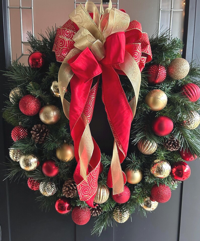 May include: A Christmas wreath with a red and gold bow, decorated with red and gold ornaments. The wreath is made of evergreen branches and has a rustic look.