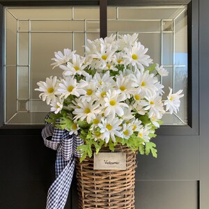 May include: A wicker basket filled with white daisies and a black and white checkered ribbon hangs on a black door with a gold "Welcome" sign. The basket has a small wooden sign that says "Welcome".