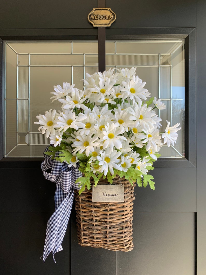 May include: A wicker basket filled with white daisies and a black and white checkered ribbon hangs on a black door with a gold "Welcome" sign. The basket has a small wooden sign that says "Welcome".