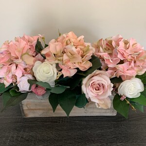 May include: A floral arrangement featuring pink hydrangeas, roses, and white flowers, accented with green leaves. The flowers are arranged in a rectangular, weathered wooden box, creating a rustic and elegant centerpiece.