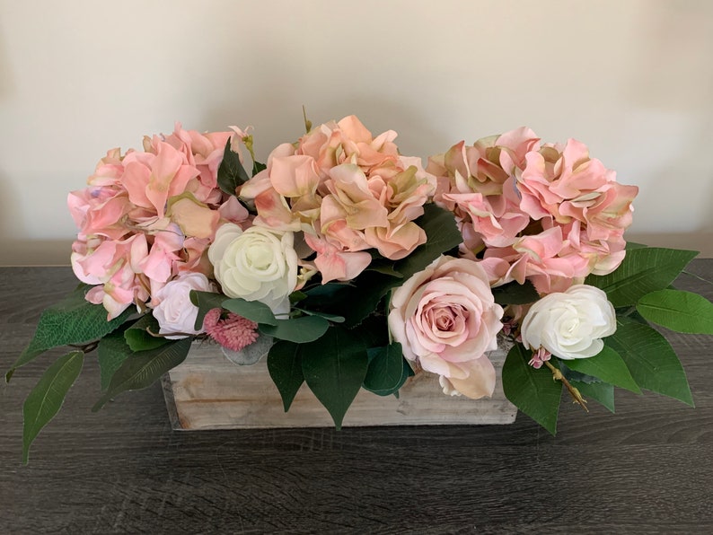 May include: A floral arrangement featuring pink hydrangeas, roses, and white flowers, accented with green leaves. The flowers are arranged in a rectangular, weathered wooden box, creating a rustic and elegant centerpiece.