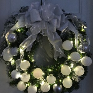 May include: A white and silver Christmas wreath with white ornaments and a white bow. The wreath is decorated with white lights and has a snowy effect.