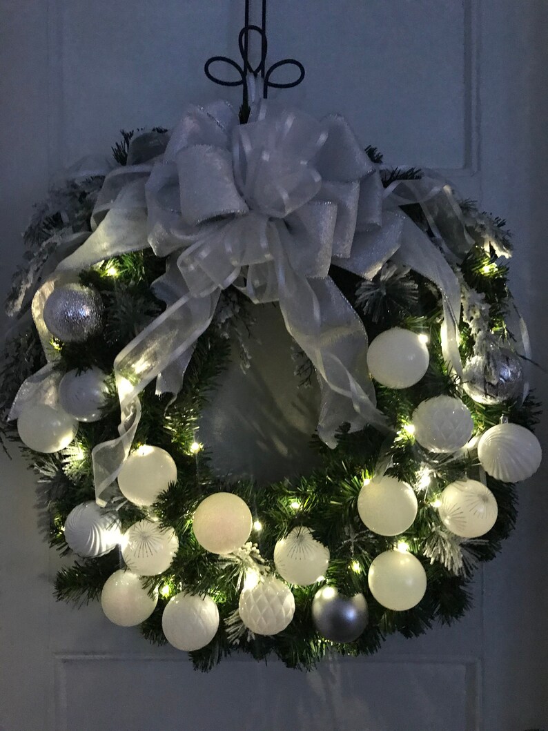 May include: A white and silver Christmas wreath with white ornaments and a white bow. The wreath is decorated with white lights and has a snowy effect.