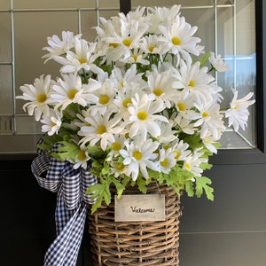 May include: A wicker basket filled with white daisies and a black and white checkered ribbon. A wooden sign with the word "Welcome" is attached to the basket.