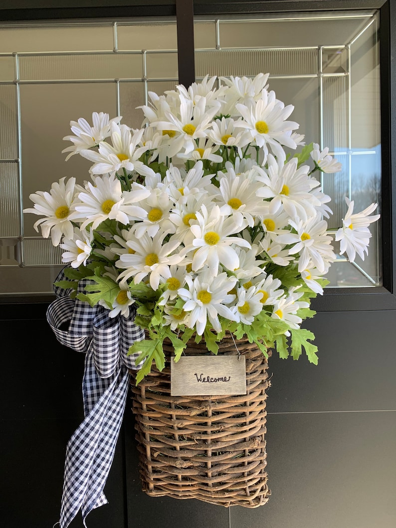 May include: A wicker basket filled with white daisies and a black and white checkered ribbon. A wooden sign with the word "Welcome" is attached to the basket.