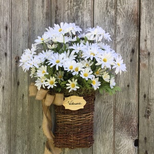 May include: A wicker basket filled with white daisies and a burlap bow hangs on a weathered wooden door. A small wooden sign with the word "Welcome" is attached to the basket.