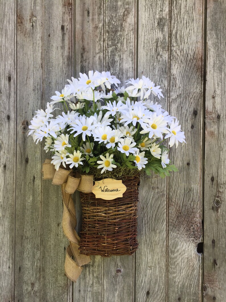 May include: A wicker basket filled with white daisies and a burlap bow hangs on a weathered wooden door. A small wooden sign with the word "Welcome" is attached to the basket.