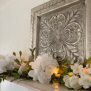 May include: A white floral garland with lights draped across a white mantel. The garland features white flowers and green leaves. A silver metal wall decor with an embossed design is above the garland.