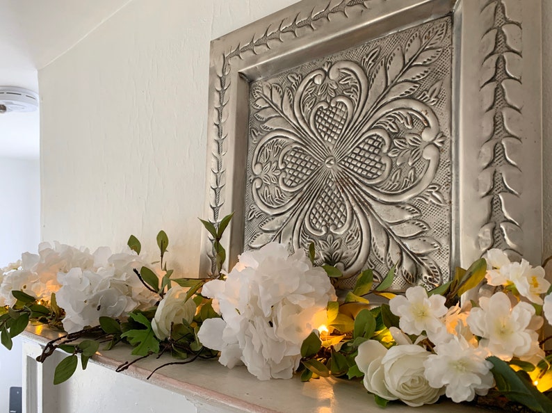 May include: A white floral garland with lights draped across a white mantel. The garland features white flowers and green leaves. A silver metal wall decor with an embossed design is above the garland.