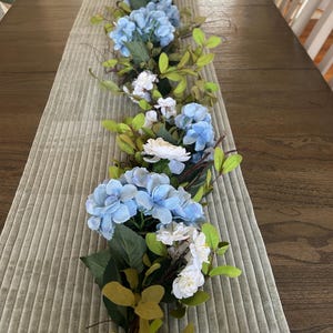 May include: A blue and white floral garland with green leaves, perfect for a table centerpiece or wedding decor.