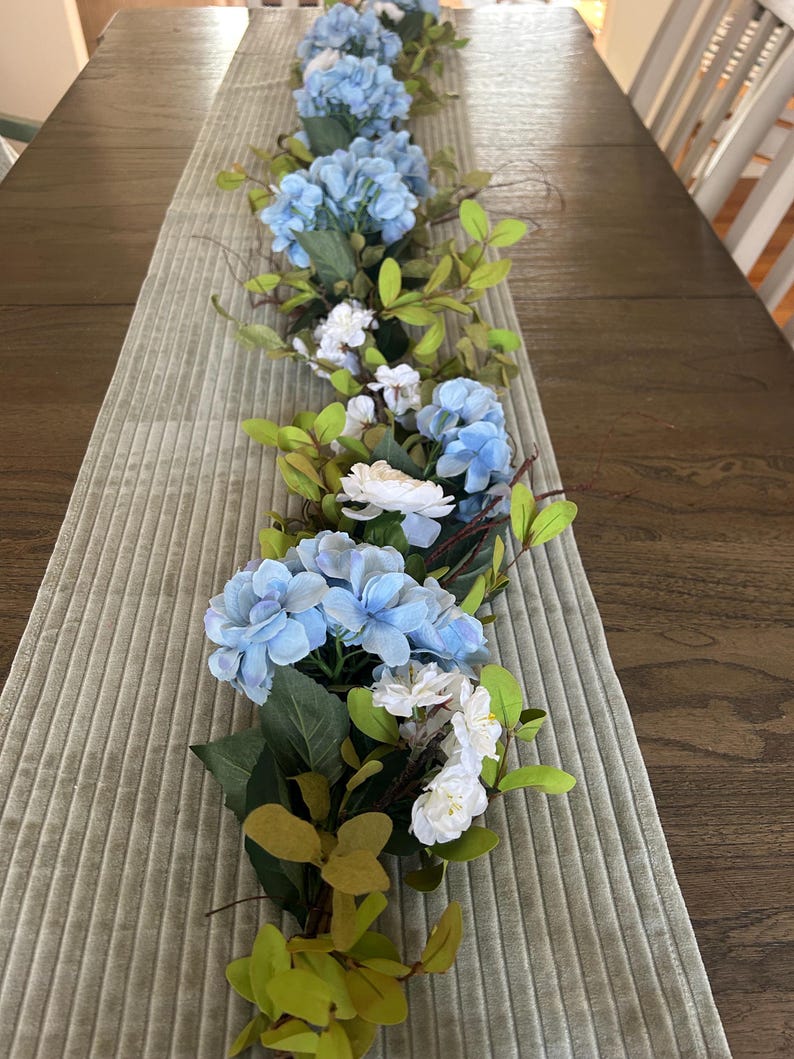 May include: A blue and white floral garland with green leaves, perfect for a table centerpiece or wedding decor.