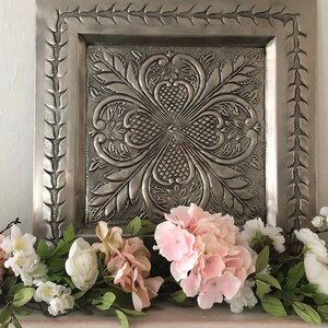 May include: A silver metal wall decor with an embossed floral design, framed with a decorative border. The decor is displayed above a garland of pink and white flowers.
