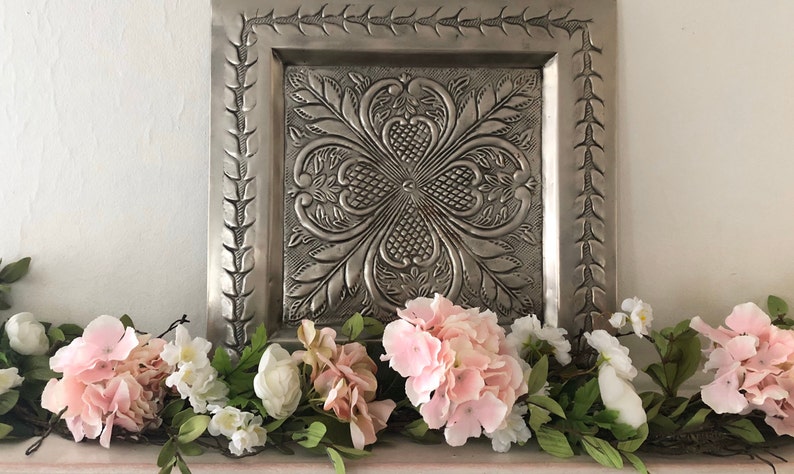 May include: A silver metal wall decor with an embossed floral design, framed with a decorative border. The decor is displayed above a garland of pink and white flowers.
