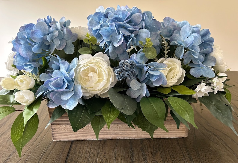 May include: A floral arrangement featuring blue hydrangeas, white roses, and greenery in a rustic wooden box. The flowers are arranged in a rectangular shape, creating a full and lush display. The color palette includes shades of blue, white, and green.