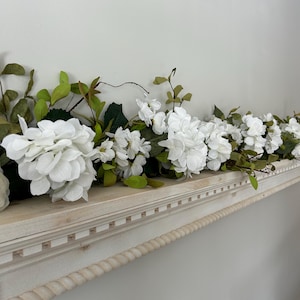 May include: White faux flower garland with green leaves draped across a white shelf with a rope trim.