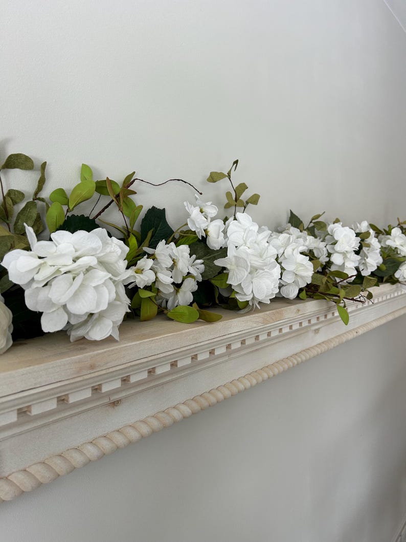 May include: White faux flower garland with green leaves draped across a white shelf with a rope trim.
