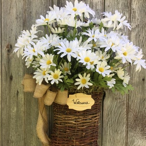 May include: A wicker basket filled with white daisies and a burlap bow hangs on a weathered wooden door. A wooden sign with the word "Welcome" is attached to the basket.