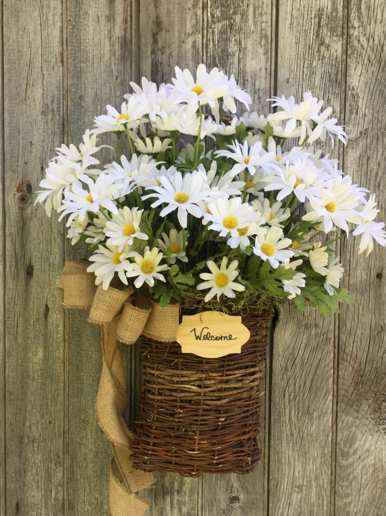 May include: A wicker basket filled with white daisies and a burlap bow hangs on a weathered wooden door. A wooden sign with the word "Welcome" is attached to the basket.