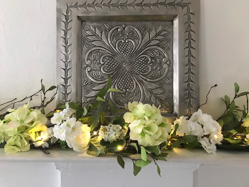 May include: A white and green floral garland with lights, decorated with white and green flowers and greenery, on a white mantel with a silver metal wall decor behind it.